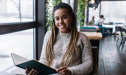 Portrait of happy African American student with braiding hairstyle holding education notepad for organisation planning and smiling at camera, cheerful hipster girl with university textbook posing