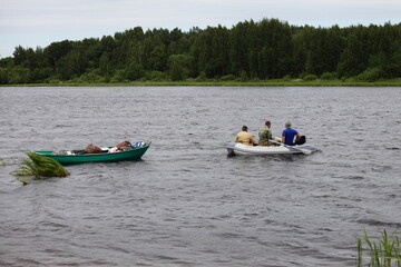 Three caucasian fishers without lifejackets floats on modern inflatable motor boat with loaded rowing boat on rope, fishing recreation on the Russian river at summer day