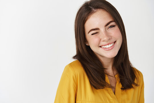 Close-up of cute and dreamy young woman smiling, winking at camera to encourage client, standing over white background