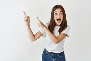 Great news hey take look. Excited happy and surprised beautiful young woman turn yout attention at banner, pointing fingers left and scream joyful, stand white background