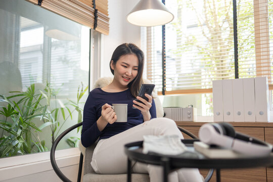 Work from home concept a confident woman sitting on a modern chair hold a cup of coffee and another hand typing a keyboard on her smart phone