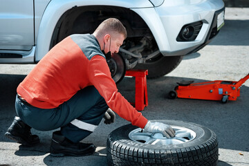 A car mechanic in a medical mask changes a wheel on a car on the street on a sunny day. Help on the...