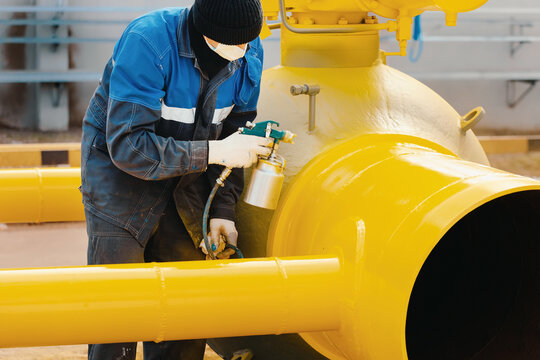A painter in working clothes paints a metal shut-off valve for gasification from a compressor gun on a summer day. Professional painting of parts. Industrial background.