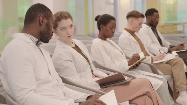 Side-view shot of multiethnic group of young male and female medical students sitting in row during lecture, whispering information to each other while taking notes