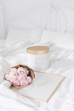 Gold gift box and white frame mockup on the bed. Bouquet of white peonies in craft packaging. Scandivanavian white interior.