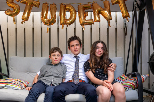 Siblings in sofa outdoors with big brother in the middle after graduation cermony, golden balloons above.