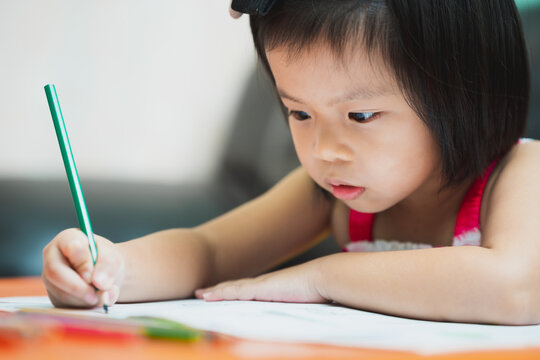 Cute Asian girl doing homework during school holidays. Children use dark green wood color to paint elaborately on their textbooks. Child age 4-5 years old. During spread coronavirus (COVID-19) disease