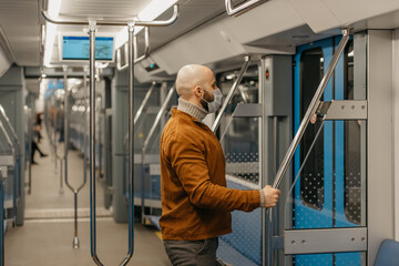 A man with a beard in a medical face mask to avoid the spread of coronavirus is preparing to leave the subway car holding the handrail. A bald guy in a mask is keeping social distance on a train.