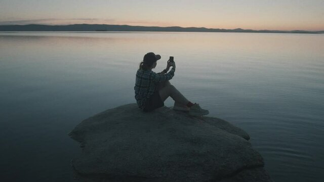 Tilt down wide shot of woman sitting on rock in lake and photographing scenic sunset with smartphone
