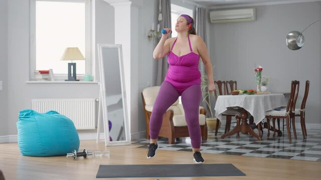 Wide shot portrait of positive obese Caucasian young woman singing dancing enjoying music in headphones exercising at home indoors. Happy plus-size lady having fun training. Aerobics and fitness