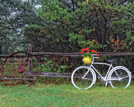 An old white painted bicycle leaning on a split rail fence with an old wagon wheel doing the same.