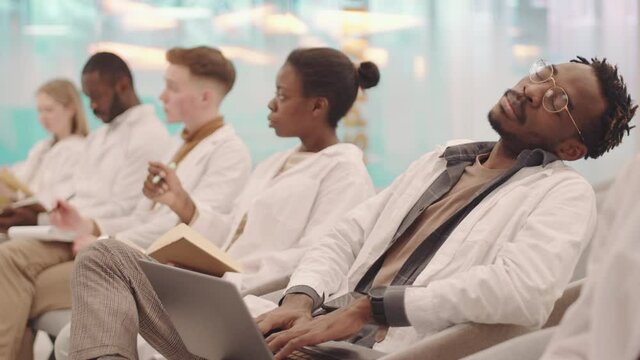 Medium shot of tired or bored young Afro American male student in white coat napping during lecture while his classmates interacting with professor asking and answering questions