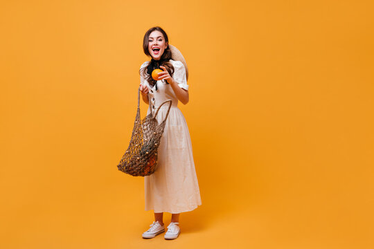 Joyful Woman Pulls Orange Out Of String Bag. Lady In White Midi Dress Posing On Orange Background