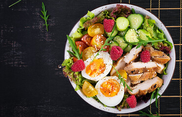 Grilled chicken meat and fresh vegetable salad of tomato, cucumber, egg, lettuce and raspberry. Ketogenic diet. Buddha bowl dish on dark background. Top view, flat lay