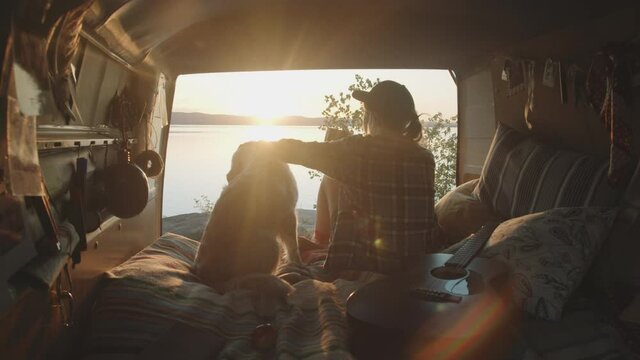 Back view of young woman sitting in camper van with opened doors, drinking tea and petting cute golden retriever dog while watching sunset over lake