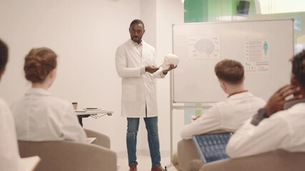 Rear view of group of young medical students sitting by desks having Anatomy lesson Young male Afro American teacher explaining brain construction pointing at posters on whiteboard and skull model - Powered by Adobe