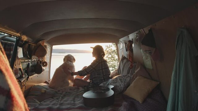Back view zoom in shot of young woman petting lovely golden retriever dog and drinking tea while sitting together in van with opened doors and watching sunset over lake