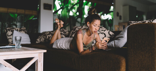 Concentrated ethnic woman using smartphone and earphones on sofa