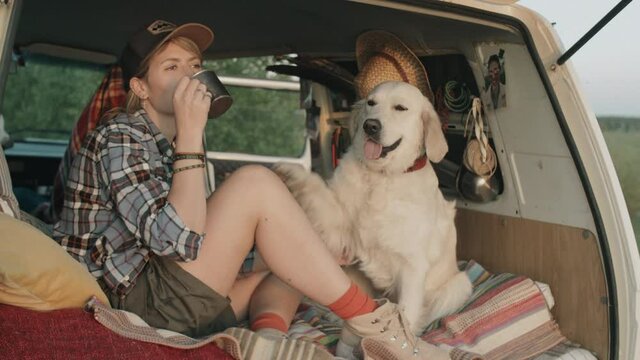 Young Caucasian woman drinking tea form steel mug and petting cute golden retriever dog while sitting together in van with opened doors in nature