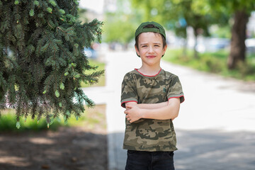 A cute boy in a T-shirt with a military print and a cap stands against the background of a green...