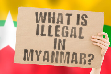 The question " What is illegal in Myanmar? " on a banner in men's hand with blurred Myanmarese flag on the background. Not allowed. Prohibitions. Outlaw. Rules. Policy. Law