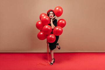 Full-length shot of charming lady wearing black midi dress and red beret. Flirtatious woman blows kiss and holds balloons