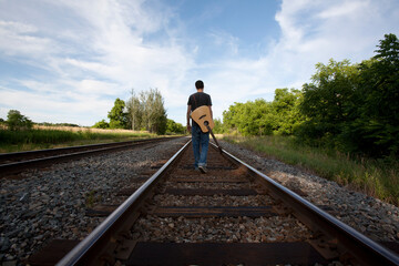 Young Man with Guitar slung on Back walking down Rail Road tracks