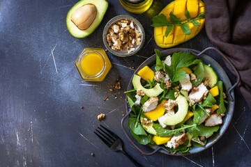 Healthy diet salad with arugula, mango, avocado, chicken grill, walnuts and vinaigrette dressing on a dark stone countertop. Top view flat lay background. Copy space.