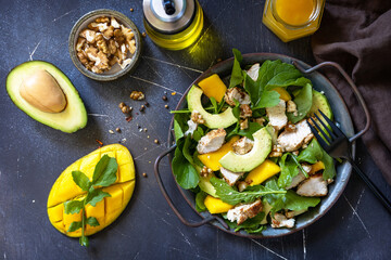 Healthy diet salad with arugula, mango, avocado, chicken grill, walnuts and vinaigrette dressing on a dark stone countertop. Top view flat lay background.