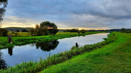 Obraz premium Grün blaue Abenddämmerung mit schmalem orangenen Streifen am Horizont. Kanal Fluss, in dem sich die Landschaft und die dunklen Wolken spiegeln. Sumpfige Sumpf Landschaft.