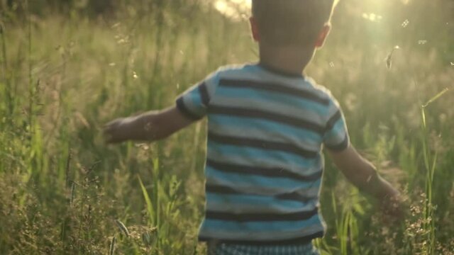 Slow motion little child boy running from camera through tall grass on summer field with beautiful sunset light. Childdream, happy childhood, freedom concept.