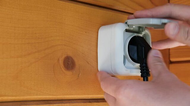 A young man inserts a black plug from an electrical appliance into an outdoor waterproof outlet with a gray cover.
