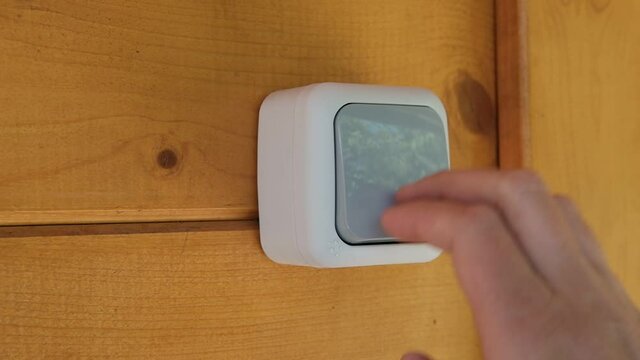A young man, with one hand, clicks on the outdoor waterproof switch several times, which is installed on a wooden wall.