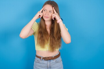 young beautiful blonde woman standing against blue background with thoughtful expression, looks away, keeps hand near face, thinks about something pleasant.