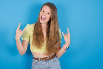 Born to rock this world. Joyful young beautiful blonde woman standing against blue background screaming out loud and showing with raised arms horns or rock gesture.