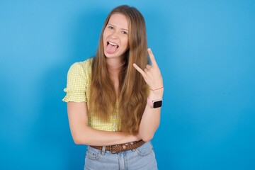 Fototapeta na wymiar Portrait of a crazy young beautiful blonde woman standing against blue background showing tongue horns up gesture, expressing excitement of being on concert of band.