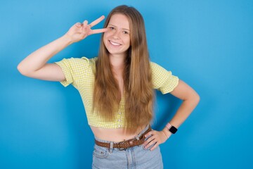 young beautiful blonde woman standing against blue background making v-sign near eyes. Leisure,...