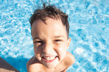 A wet child with a funny, emotional face in a pool of blue water. The kid grimaces, has fun in the...
