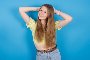 Obraz na płótnie Canvas young beautiful blonde woman standing against blue background relaxing and stretching, arms and hands behind head and neck smiling happy
