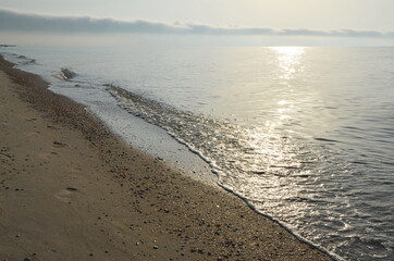 Seaside, sandy shore, with small waves.
 Morning sunrise.
The sun is reflected in the water.
There are small white clouds on the horizon.