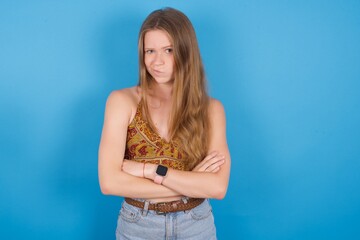 Picture of angry young beautiful blonde woman standing against blue background crossing arms. Looking at camera with disappointed expression.
