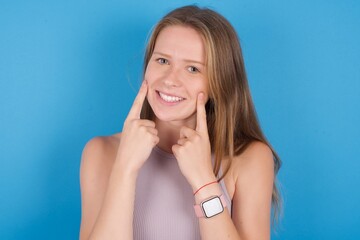 Happy young beautiful blonde woman standing against blue background with toothy smile, keeps index fingers near mouth, fingers pointing and forcing cheerful smile