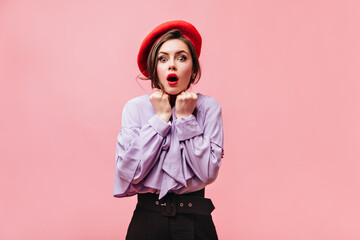 Green-eyed brunette woman in bright beret and blouse with fright looks into camera on pink background