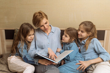 a happy mother with three daughters reads a fairy tale book before going to bed from the bedroom. happy family, reading a book aloud to children, the process of teaching and raising children