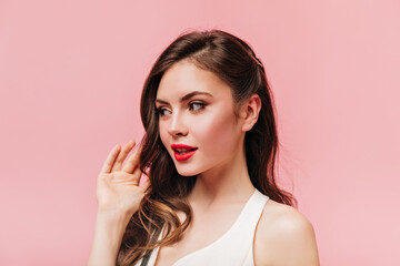 Lady in white top touches her wavy hair. Shot of brunette woman with red lips on pink background