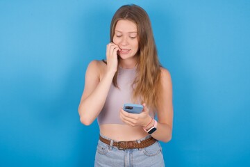 Portrait of pretty frightened young beautiful blonde woman standing against blue background chatting biting nails after reading some scary news on her smartphone.