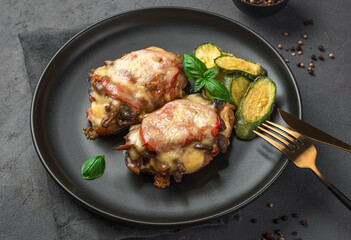 Baked meat with mushrooms, tomatoes and cheese with a side dish of their zucchini on a dark background.