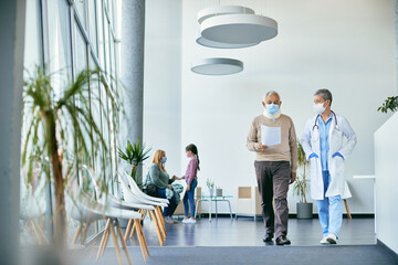 Senior man with neck injury and his doctor wearing face masks while walking through hallway and talking at medical clinic.