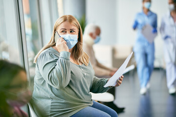 Woman wearing face mask while sitting at hospital waiting room and making a phone call.