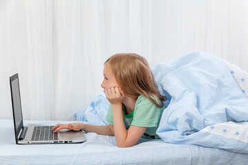 The girl lies at home on the bed under a blanket with a laptop. Distance learning. Computer games. Lessons. homework. education.